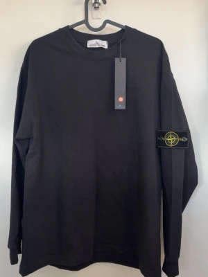 Stone Island tröja - Svart långärmad tröja från Stone Island med klassisk rund hals och ikonisk patch med logga på vänster ärm. Tillverkad i mjuk bomull för en clean och stilren look. Perfekt för dig som gillar streetwear och vill ha något enkelt men ändå exklusivt.