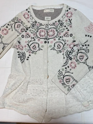 Odd Molly kofta med spets och mönster - Supersöt ljusgrå kofta från Odd Molly med broderade blommor i rosa och svart på bröst och ärmar. Koftan har rund halsringning, knappar framtill och vackra spetsdetaljer nedtill. Perfekt för dig som gillar romantisk och bohemisk stil.