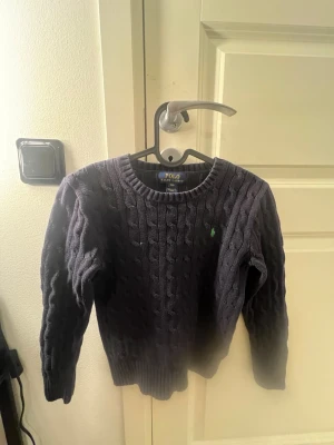 Mörkblå kabelstickad tröja Polo Ralph Lauren - Mörkblå kabelstickad tröja från Polo Ralph Lauren med rund halsringning och lång ärm. Tröjan har det klassiska gröna Polo-logot på bröstet och ribbstickade muddar vid ärmslut och nederkant. Perfekt för en stilren och avslappnad look.