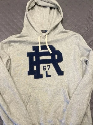 Grå hoodie från Polo Ralph Lauren - Snygg grå hoodie från Polo Ralph Lauren med stor mörkblå PR 67-logga på bröstet. Hoodien har huva med vita snören och en klassisk känguruficka framtill. Materialet är mjukt och bekvämt, perfekt för en avslappnad look.