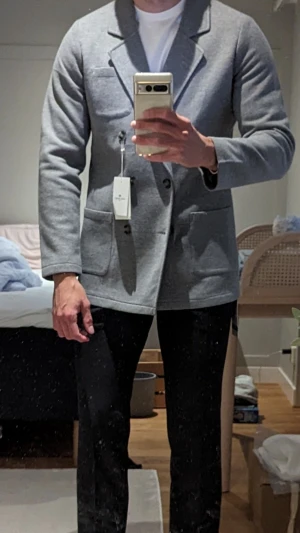 Grå dubbelknäppt kavaj från John Henric - Snygg grå stickad kavaj med dubbelknäppning och breda slag. Kavajen har två stora framfickor och en bröstficka. Tillverkad i ett mjukt, stickat material som ger en avslappnad men stilren look. Perfekt att bära över en t-shirt för en modern vibe.