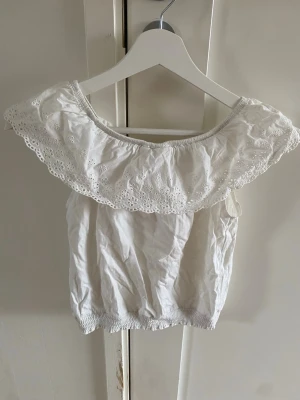 Vit offshoulder topp med spetsvolang - Söt vit offshoulder topp med bred spetsvolang runt axlarna och smockad nederkant. Toppen är ärmlös och har en romantisk känsla med broderade detaljer i volangen. Perfekt för varma dagar och somriga outfits.
