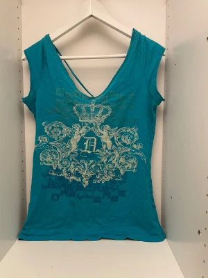 Turkos topp med tryck och öppen rygg - Snygg turkos topp med stort dekorativt tryck framtill och djup v-ringning både fram och bak. Toppen har tunna axelband som korsas i ryggen för en cool detalj. Perfekt för dig som vill sticka ut lite extra. Äkta DKNY. Tröjan har en lite slit fläck nedre vänster sida.