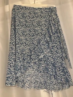 Blommig omlottkjol i blått och vitt🌟 - NY Söt omlottkjol med volangkant i blått och vitt blommönster. Kjolen har knytband i sidan och är tillverkad i ett lätt, luftigt material som passar perfekt till sommaren. 🌸✨🌟