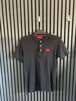 Svart pikétröja från Hugo Boss - Snygg svart pikétröja från Hugo Boss med klassisk krage och knappar framtill. Tröjan har korta ärmar och en röd logotyp på bröstet samt röd detalj på insidan av kragen. Perfekt för en stilren look.