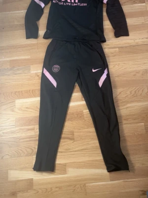 Svart PSG träningsset från Nike - Säljer ett svart träningsset från Nike och PSG med rosa detaljer. Setet består av en långärmad tröja och matchande byxor. Tröjan har tryckt PSG-logga och texten 'Accor Live Limitless' på bröstet. Byxorna har smal passform, rosa detaljer och Nike-logga.