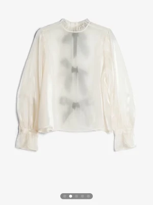 Vit transparent blus med svarta rosetter - Stilren vit transparent blus i glansigt material med lång ärm och volangkrage. Baksidan har tre stora svarta rosetter som knyts, vilket ger en unik och trendig look. Perfekt för dig som vill sticka ut och gillar detaljer.