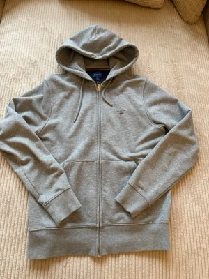 Grå zip hoodie från Gant - Snygg grå hoodie från Gant med dragkedja framtill och justerbar huva med snören. Klassisk passform, ribbade muddar och fickor på magen. Liten Gant-logga broderad på bröstet. Perfekt för en avslappnad och stilren look.