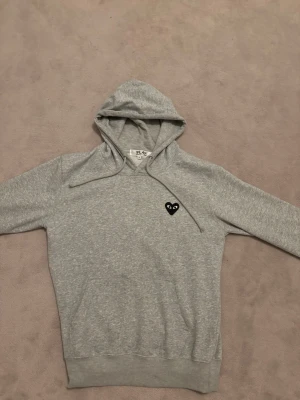 Grå hoodie från Comme des Garçons Play - Säljer en grå hoodie från Comme des Garçons Play med det ikoniska svarta hjärta-ögon-loggan på bröstet. Hoodien är i toppskick me inga defekter, använd ett fåtal gånger och säljer för ett mycket bra pris. Pris går såklart att diskutera, skriv vid funderingar!