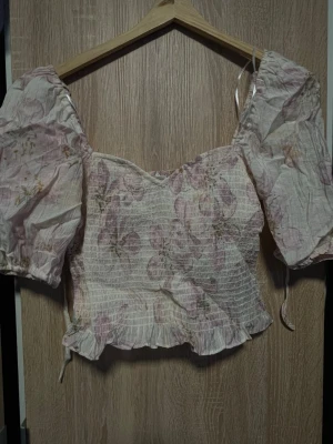 Blommig puffärm topp från H&M - Söt topp från H&M med puffärmar och smockad kropp. Tunt, krämvitt tyg med ljusrosa och beige blommönster. Kortare modell med volangkant nedtill och band vid ärmarna. Perfekt för en romantisk och trendig look. Aldrig använt 😊