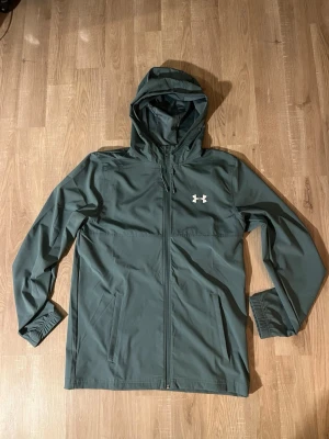 Under Armour Windbreaker Blå/Turqoise - Snygg Turqoise/Ljusblå vindjacka från Under Armour med huva och dragkedja framtill. Jackan har två sidofickor och logga på bröstet samt stor text på ryggen. Köptes i Storbritannien när jag var där under nyår detta året. 