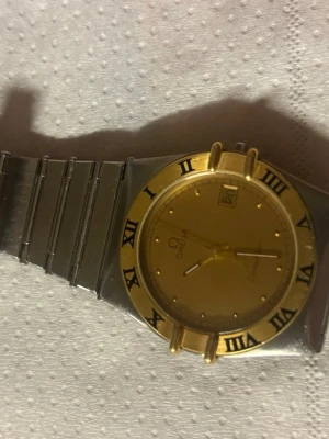 Omega guldfärgad klocka med datum - Stilren Omega klocka med guldfärgad urtavla och bezel, romerska siffror och datumvisning. Armbandet är i rostfritt stål och klockan har klassisk design som passar till många stilar. Perfekt accessoar för dig som gillar exklusiva detaljer.