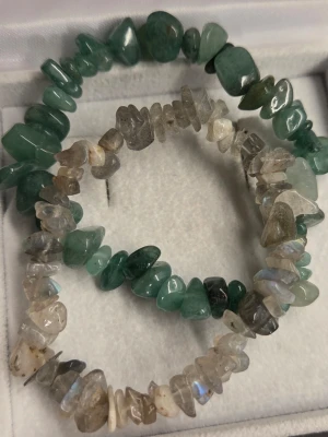 Stenarmband med ädelstenar i grönt och grått - Två elastiska armband med oregelbundna ädelstenar av gröna och grå stenar. De gröna stenarna har en djup, naturlig ton medan de grå skimrar lätt i ljuset. Perfekt för dig som gillar naturliga material och en bohemisk stil. Justerbara armband.