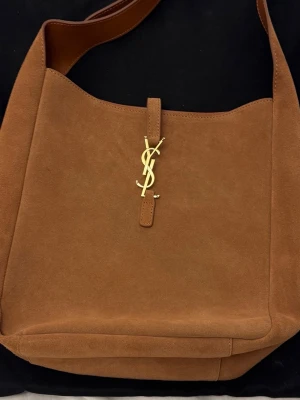 Äkta mocka handväska från Saint Laurent (le 5 a 7) - Säljer en snygg handväska i äkta brun mocka från Saint Laurent med ikoniskt YSL-emblem i guld framtill. Perfekt skick, ingen lukt, helt oanvänt. Äkta kostar ungefär 28-30 tusen.