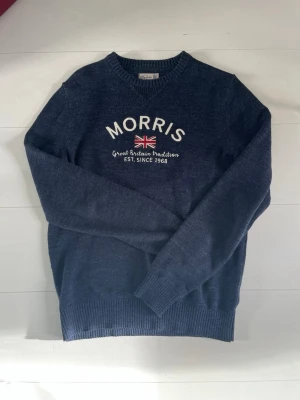 Marinblå Morris Tröja - Snygg mörkblå stickad tröja från Morris med broderad Union Jack-flagga och texten 'Great Britain tradition EST. SINCE 1968' på bröstet. Tröjan har rund halsringning och ribbade muddar vid ärmslut och nederkant. Perfekt för en klassisk och avslappnad stil.