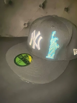 New Era Yankees svart keps 59Fifty - Svart New Era 59Fifty keps med broderad vit Yankees-logga och blå Frihetsgudinnan framtill. Baksidan har ett rött äpple och MLB-logga. Klassisk flat brim och broderade detaljer. Perfekt för dig som gillar streetwear och sportig stil.