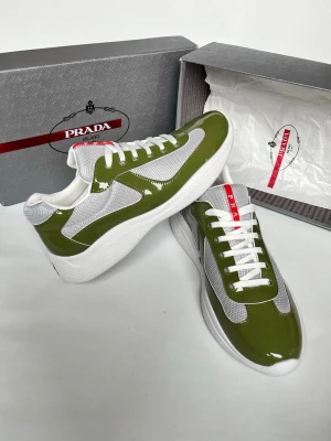 Prada gröna och vita sneakers - Snygga sneakers från Prada i blankt grönt och vitt med meshpaneler för extra ventilation. Skorna har vita snören, tjock vit sula och röda detaljer på plösen. Perfekta för dig som vill ha en trendig och exklusiv look.