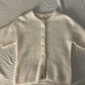 Vit ribbad kofta från Gina Tricot - Mysig vit kofta från Gina Tricot med ribbad struktur och rund halsringning. Koftan har korta ärmar och knäpps med pärlemorskimrande knappar framtill. Perfekt att slänga över en topp för en enkel och stilren look.