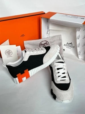 Hermès svartvita sneakers med orange H - Snygga sneakers från Hermès i svart mesh och vitt mocka med en stor orange H-detalj på sidan. Skorna har vita snören och en chunky, räfflad sula i vitt. Perfekt för dig som gillar exklusiva och trendiga skor med unika detaljer.