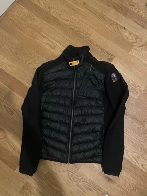 Svart dunjacka från Parajumpers - Snygg svart dunjacka från Parajumpers med mjuka stickade ärmar. Dragkedja framtill, hög krage med gul detalj och logga på ärmen. Perfekt för lager-på-lager och streetwear-vibe. Skriv om ni har några frågor ✌️