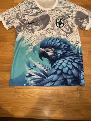 Nike Brasil CBF t-shirt med blå papegoja - Unik sporttröja från Nike med Brasil CBF-logga. T-shirten har ett färgstarkt tryck med en stor blå papegoja och detaljer i vitt, grått och turkos. Tillverkad i ett lätt och ventilerande material, perfekt för träning eller streetwear. Rund halsringning och korta ärmar.