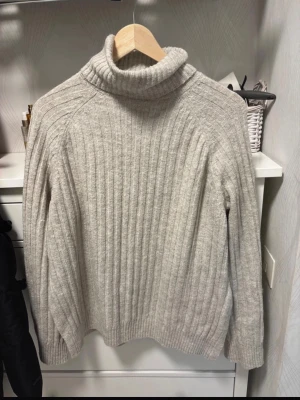 Beige ribbstickad polotröja - Mysig beige ribbstickad polotröja med hög krage och långa ärmar. Tröjan har en avslappnad passform och är tillverkad i ett mjukt, tjockt material som passar perfekt för kyliga dagar. Snygg detalj med slits i sidan.