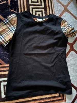 Svart Burberry t-shirt med rutiga ärmar - Snygg svart t-shirt från Burberry med klassiska beige, röda och svarta rutiga kortärmar. T-shirten har en rund halsringning och är tillverkad i mjuk bomull. Perfekt för dig som vill ha en unik twist på en basic tee.