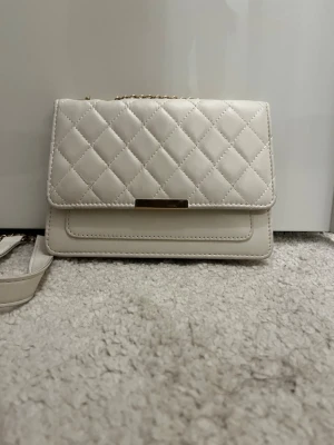 Beige quiltad axelväska med guldkedja - Säljer en stilren beige axelväska med quiltat mönster och guldfärgad kedja som axelrem. Väskan har en rektangulär form och ett elegant metallspänne framtill. Perfekt för dig som gillar klassisk och trendig design.