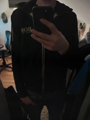 Svart hoodie från Hugo Boss - Säljer en svart hoodie från Hugo Boss med gulddetaljer och dragkedja framtill. Stora BOSS-tryck på bröstet och längs dragkedjan. Hoodien har huva och är tillverkad i ett mjukt material som känns skönt mot huden. Perfekt för dig som vill ha en stilren och exklusiv look. Inga skador osv. 