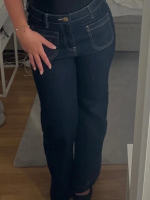 Mörkblå utsvängda jeans med kontrastsömmar - Säljer ett par mörkblå jeans med utsvängda ben och tydliga kontrastsömmar. Jeansen har höga fickor fram och klassisk knappgylf.