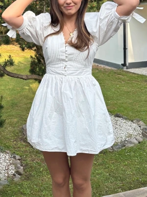 Vit kort klänning med puffärmar - Superfin vit kort klänning med puffärmar och knappar framtill. Klänningen har v-ringning, markerad midja och knytband vid ärmsluten. Tillverkad i ett lätt bomullstyg som känns luftigt och fräscht.