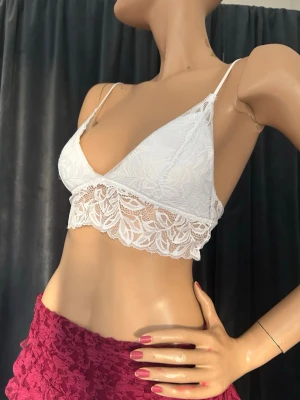 Vit spetslinnetopp med tunna band - Säljer en vit bralette i spets med tunna axelband och triangelformade kupor. Nedre kanten har ett bredare spetsband med blommigt mönster som ger en romantisk look. Använd fåtal gånger♥️