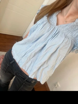 Ljusblå blus med volangdetaljer - Söt ljusblå blus med volangdetaljer vid halsringningen och korta ärmar. Blusen har en lös passform och är tillverkad i ett tunt, luftigt material som passar perfekt till varma dagar. Enkel att styla med både jeans och kjol. Storlek 170/s