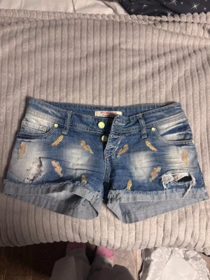 Blå denimshorts med broderade fjädrar - Snygga blå jeansshorts med slitningar och broderade fjädrar i gult och orange. Shortsen har upprullade benslut, fem fickor och dubbla knappar framtill. Perfekta för en avslappnad och trendig look.
