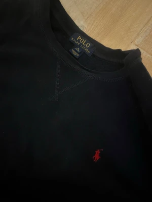 Svart sweatshirt från Polo Ralph Lauren - Klassisk svart sweatshirt från Polo Ralph Lauren med rund halsringning och den ikoniska röda logotypen broderad på bröstet. Tröjan har ribbade muddar vid ärmslut och nederkant. Perfekt för en clean och stilren look.