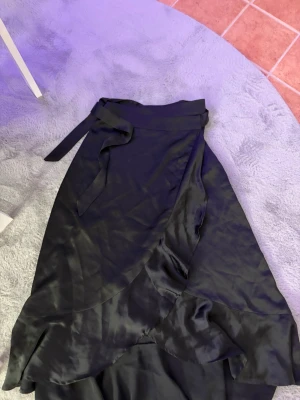 Svart omlottkjol från Object - Säljer en svart omlottkjol från Object i blankt material. Kjolen har volangdetaljer och knyts i midjan för en snygg passform. Perfekt för dig som gillar stilrena plagg med en twist. Endast använd en gång.