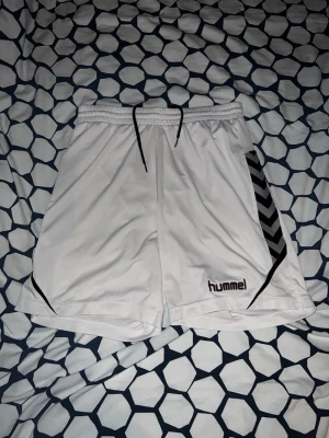 Vita träningsshorts från Hummel - Säljer ett par vita träningsshorts från Hummel med svart snörning i midjan och svarta detaljer på sidorna. Shortsen har det klassiska Hummel-mönstret längs ena benet och är tillverkade i ett lätt och snabbtorkande material, perfekta för sport och träning.