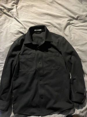 Svart overshirt från Zara - Stilren svart overshirt från Zara med två stora bröstfickor och knappar framtill. Klassisk krage och raka ärmar ger en clean look. Perfekt att bära öppen eller stängd över en t-shirt. Materialet känns mjukt och har en lätt struktur.