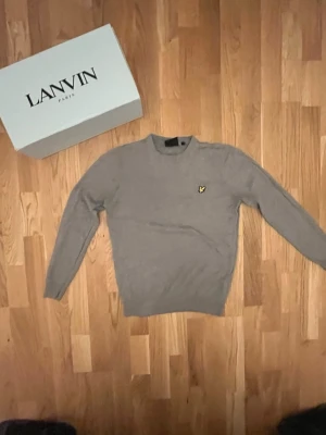 Beige stickad tröja från Lyle & Scott - Säljer en stilren beige stickad tröja från Lyle & Scott med rund halsringning och den klassiska loggan broderad på bröstet. Tröjan har långa ärmar och ribbade muddar vid ärmslut och nederkant. Perfekt för en clean och avslappnad look.