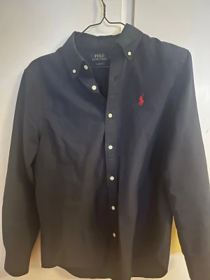 Marinblå skjorta från Polo Ralph Lauren - Klassisk marinblå skjorta från Polo Ralph Lauren med button-down krage och vita knappar. Skjortan har lång ärm och den ikoniska röda logotypen broderad på bröstet. Tillverkad i mjuk bomull för en bekväm känsla.