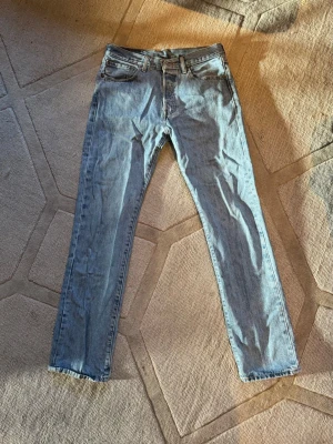 Levi's 501 ljusblå raka jeans - Klassiska Levi's 501 jeans i ljusblå denim med rak passform och knappgylf. Tidlös modell som funkar till allt och har den ikoniska läderlappen bak i midjan. Perfekta för dig som gillar vintagevibbar och avslappnad stil.