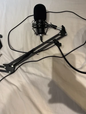 Svart mikrofon med stativ och arm - Säljer en svart mikrofon med puffskydd, monterad i ett metallstativ med justerbar arm. Perfekt för streaming, podd eller gaming. Mikrofonen har en klassisk rund form och ansluts med kabel. Armen är i svart metall och kan fästas på skrivbord. 