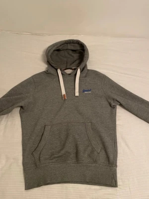 Grå hoodie från Superdry - Snygg grå hoodie från Superdry med klassisk känguruficka och vita dragsnören i huvan. Liten blå Superdry-logga på bröstet. Tillverkad i mjukt sweatshirtmaterial som är perfekt för chill dagar. Passar dig som gillar enkel och stilren streetwear. Pris kan diskuteras 