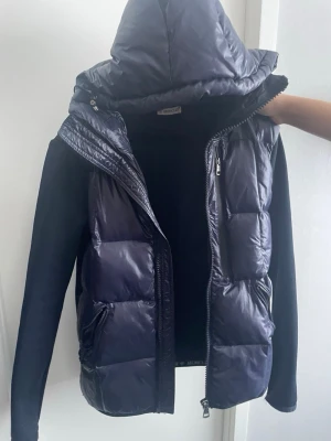 Moncler cardigan blå  - Snygg mörkblå pufferjacka från Moncler med glansig finish och vadderade paneler. Jackan har huva, dragkedja framtill och två fickor med dragkedja. Ärmarna är i ett mjukare tyg som kontrasterar mot den quiltade kroppen. Perfekt för kalla dagar.