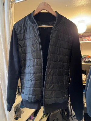 Svart quiltad cardigan från Barbour - Svart cardiganjacka från Barbour International med quiltad front och mjuka stickade ärmar. Jackan har ribbade muddar, dragkedja framtill och en diskret logopatch på ärmen. Perfekt för lager-på-lager och en clean look.