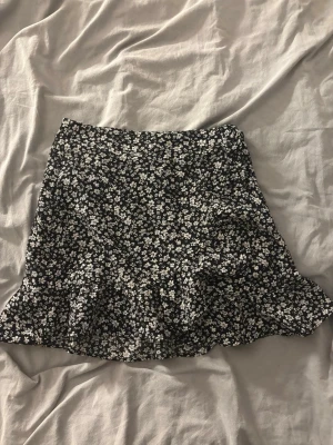 Svart blommig kortkjol med volang - En svart kortkjol med vita blommor och volang längst ner. Kjolen har en lätt utsvängd form och är perfekt för dig som gillar en söt och trendig stil. Materialet känns mjukt och lätt, perfekt för varmare dagar.