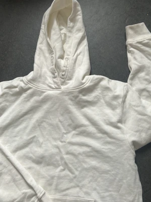 Vit hoodie med magficka - Klassisk vit hoodie med huva och magficka. Tillverkad i mjukt bomullstyg och har ribbade muddar vid ärmslut och nederkant. Perfekt för en avslappnad och stilren look.