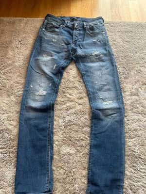 Jack and Jones Jeans Ripped - Säljer ett par blå jeans med slitningar på framsidan. Jeansen är slim fit, glenn Model med klassisk femficksmodell. Perfekta för en avslappnad och trendig look. Storlek 27/32