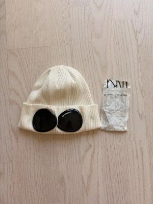 Vit mössa med goggles från C.P. Company - Säljer en ribbstickad vit mössa från C.P. Company med de ikoniska svarta goggles-linserna framtill. Mössan är tillverkad i mjukt material och har en uppvikt kant. Perfekt accessoar för dig som vill sticka ut och addera streetwear-vibe till din outfit.