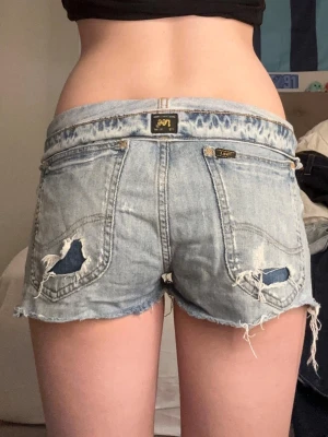 Lågmidjade jeansshorts från Lee - Säljer ett par ljusblå, lågmidjade jeansshorts från Lee med slitna detaljer och fransig kant. Shortsen har klassiska bakfickor och en avslappnad, loose passform. Perfekta för sommaren och ger en cool, avslappnad vibe. 40cm raktöver midjan❤️